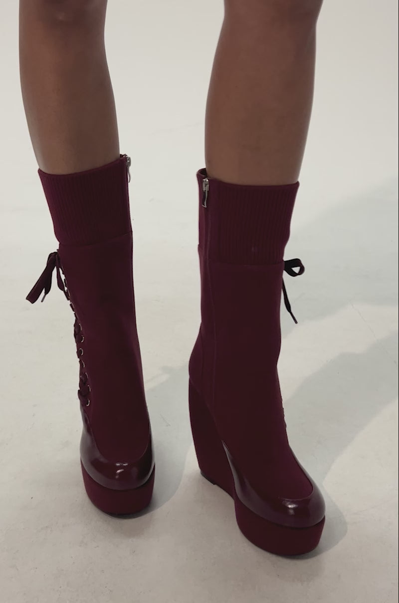 Video of woman modeling the AKIRA GOLD WHATEVER BURGUNDY CANVAS WEDGE BOOTIE— boots with wedge heels, side zippers, lace-up detail on one boot, ribbed knit cuffs, and glossy toe caps