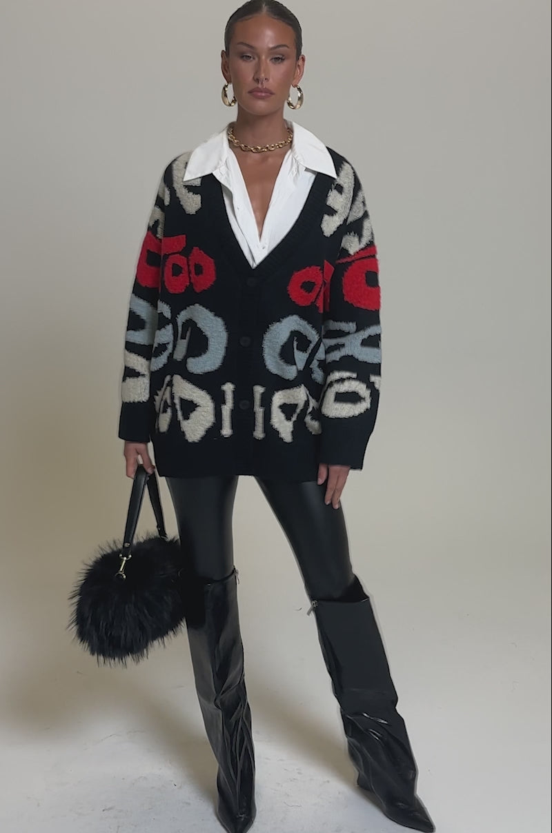Video of woman modeling the GUESS WHO CARDIGAN, featuring bold red, white, and gray abstract patterns, over a white collared shirt and black leather pants. She finishes the outfit with a black faux fur accessory and gold hoop earrings.