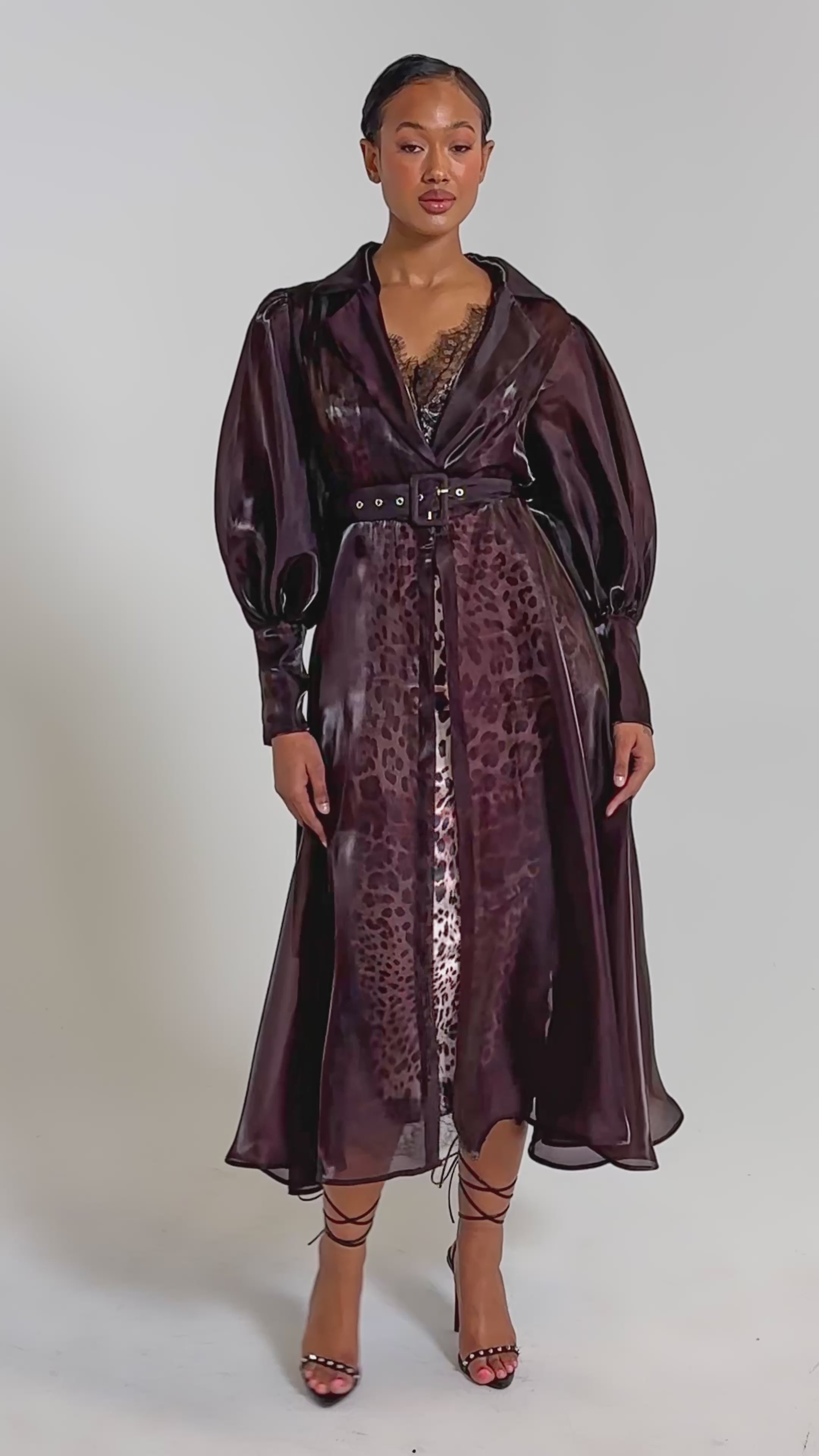 A video of a woman faces forward in the PRETTY LIL THING ORGANZA TRENCH, a sheer brown belted coat layered over a leopard-print slip. 