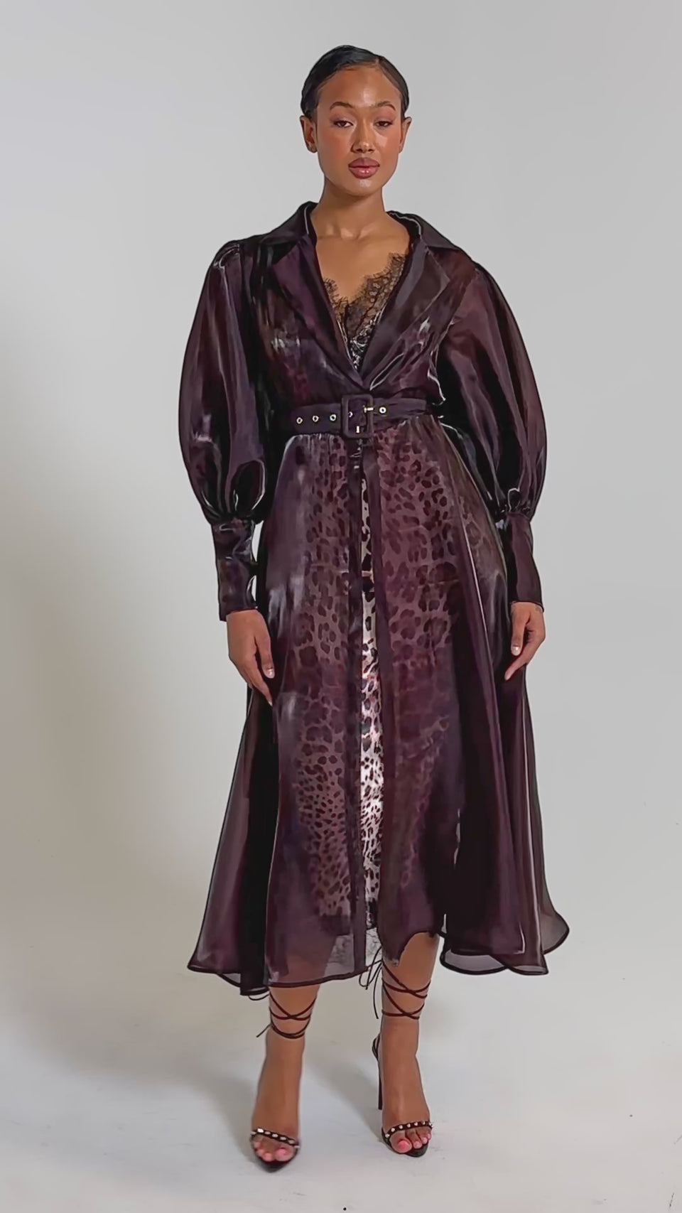A video of a woman faces forward in the PRETTY LIL THING ORGANZA TRENCH, a sheer brown belted coat layered over a leopard-print slip. 