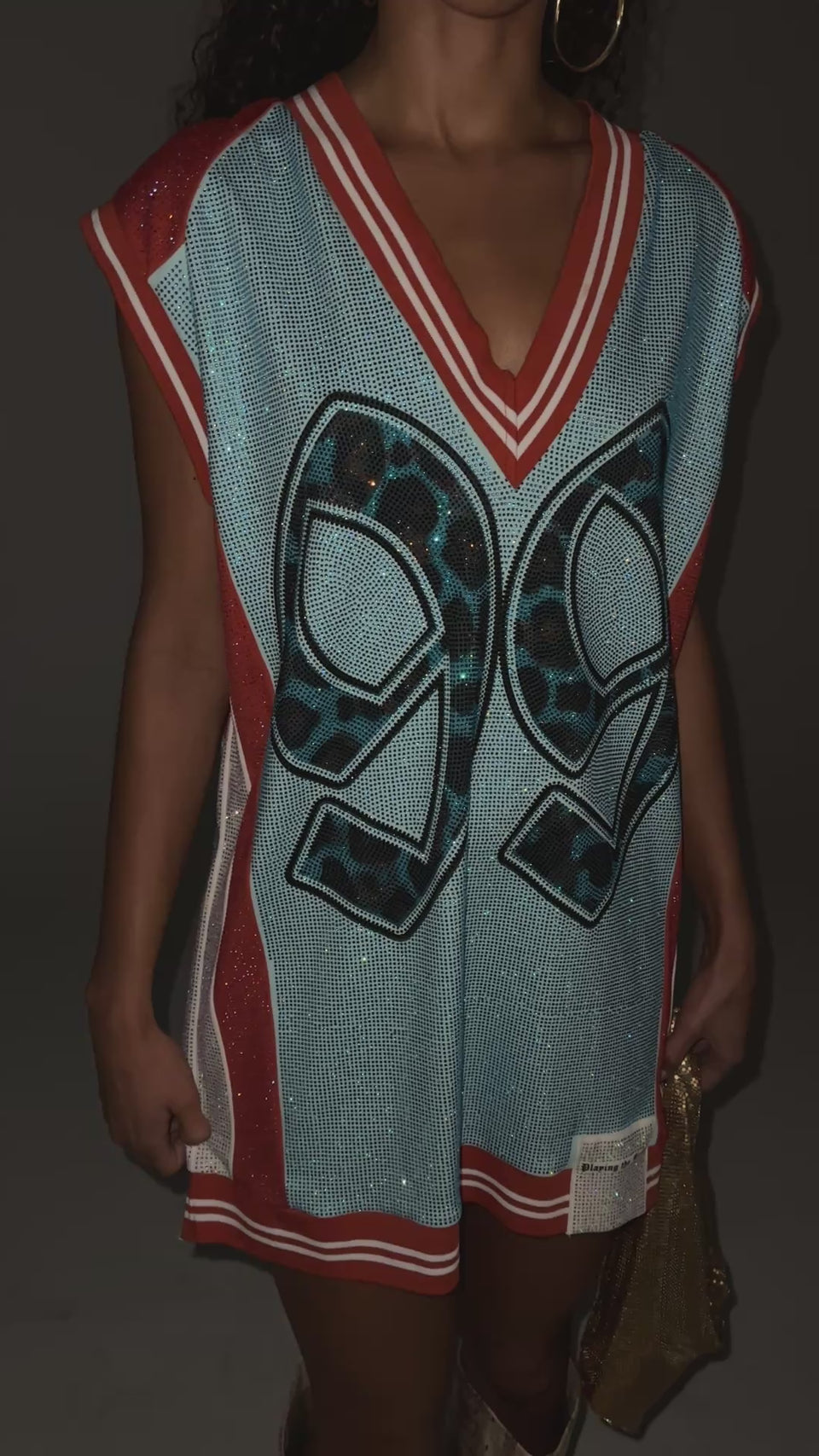 A video of a model wearing the RYAN COLORBLOCK SLEEVELESS JERSEY IN BLUE COMBO. A bedazzled colorblock jersey dress made to bring sporty glam energy to your going-out lineup. Featuring striped ribbed knit trim on the hemlines, a V-neckline, a sleeveless design, a bold “99” graphic on the front and back, contrasting side panels, and glittering rhinestone embellishments throughout.