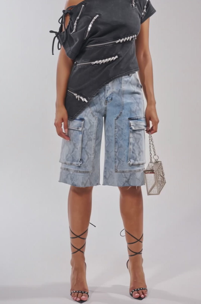 A video of a woman wearing the ROGUE ZIPPER ASYMMETRIC TEE with zipper detail, light blue knee-length cargo shorts, strappy black heels, oversized sunglasses, and carrying a small metallic handbag.