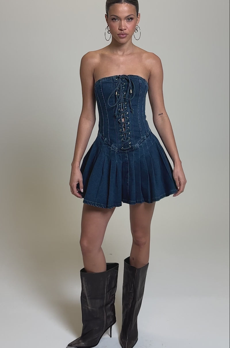 Video of woman modeling the LACE ME UP DENIM MINI DRESS, a strapless dark blue denim mini with a lace-up panel and pleated skirt. She accessorizes with hoop earrings, has slicked-back hair, and poses against a plain background.