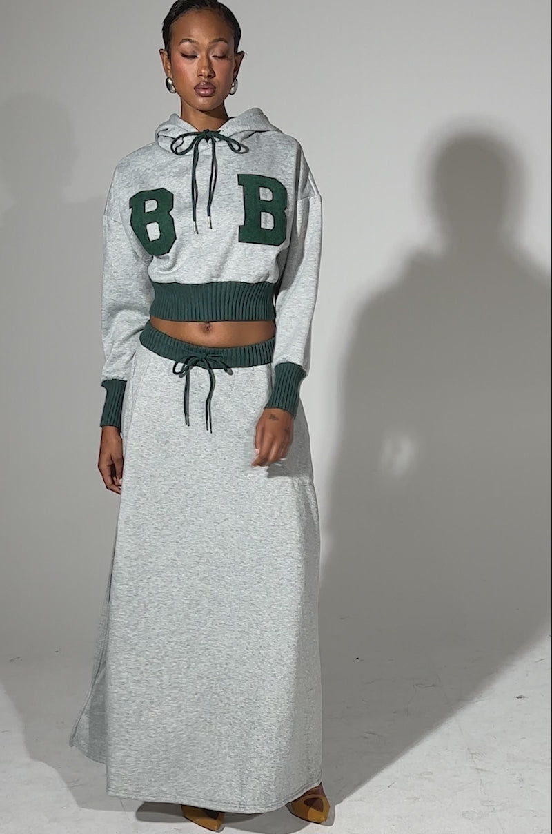 A video of a woman wearing the COLLEGIATE FRENCH TERRY HOODED SWEATSHIRT—gray with green cuffs and bold "B B" letters—paired with a matching long gray skirt and tan heels against a light background.