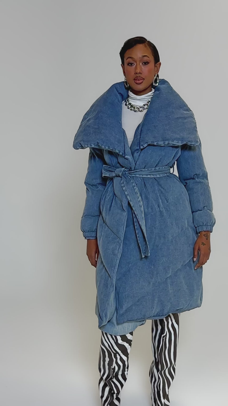 Video of woman modeling the I'M OUTSIDE DENIM PUFFER, a long light blue puffer coat with quilted padding and an oversized shawl collar, paired with a white turtleneck and chunky chain necklace against a plain background.