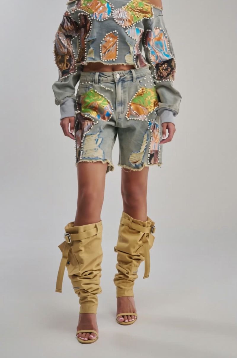 Video of woman modeling the PATCHED UP LOW RISE BROCADE BERMUDA with an off-shoulder, embellished denim top. She pairs this chic look with tan slouchy knee-high boots and gold sandals, standing confidently against a light background.
