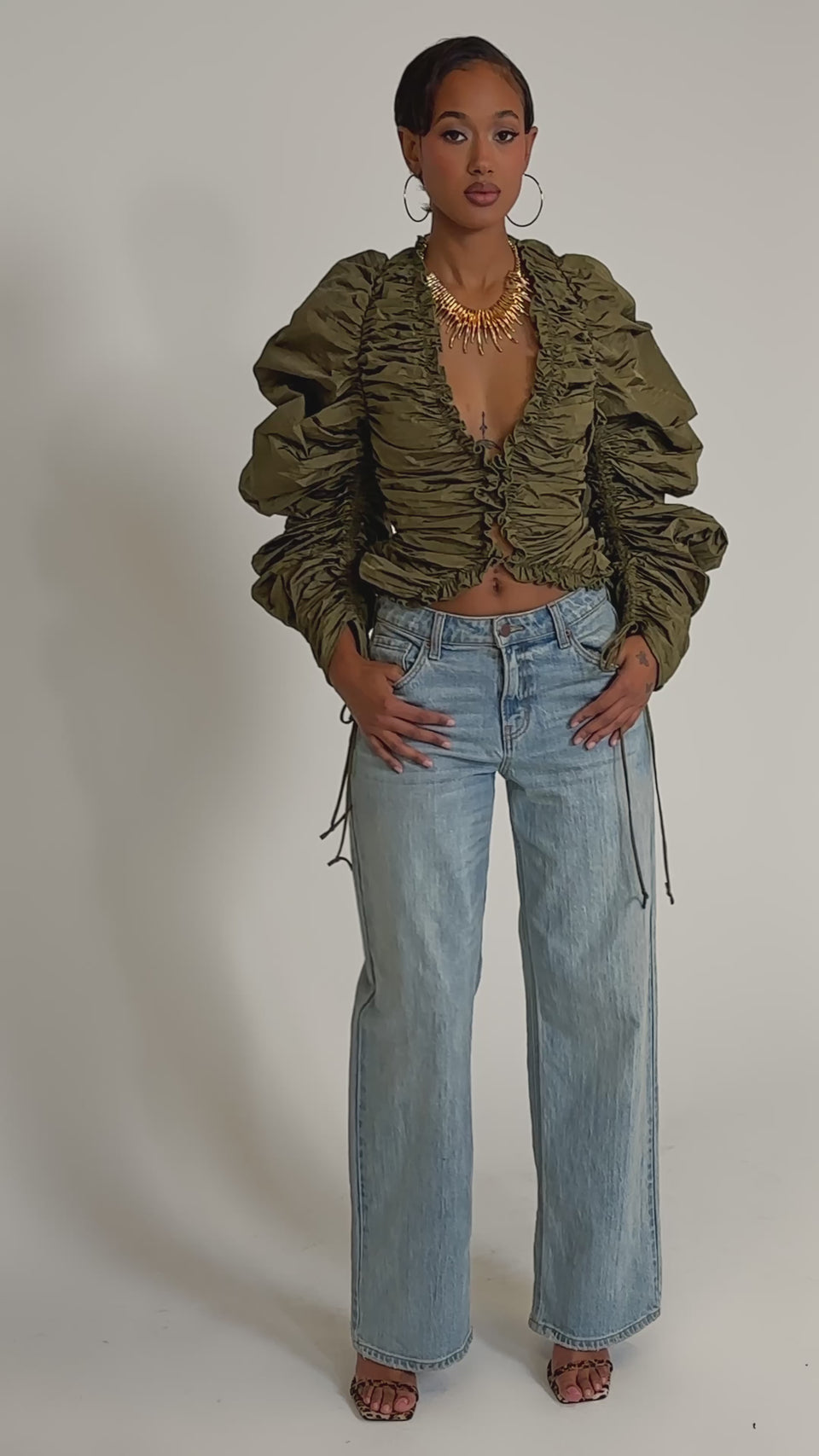 A video of a woman in the IN IT TO WIN IT RUCHED BLOUSE, a metallic olive green top with adjustable drawstring sleeves, paired with light blue wide-leg jeans, heeled sandals, large hoop earrings, and a statement necklace on a plain white background.