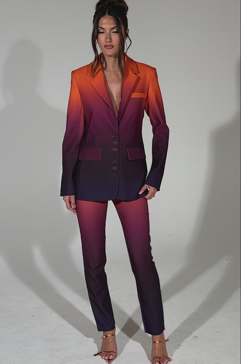 A video of a woman posing confidently in the RISE AND SHINE OMBRE BLAZER and matching pantsuit, featuring orange, pink, and purple hues with strappy heels, against a light gray background.