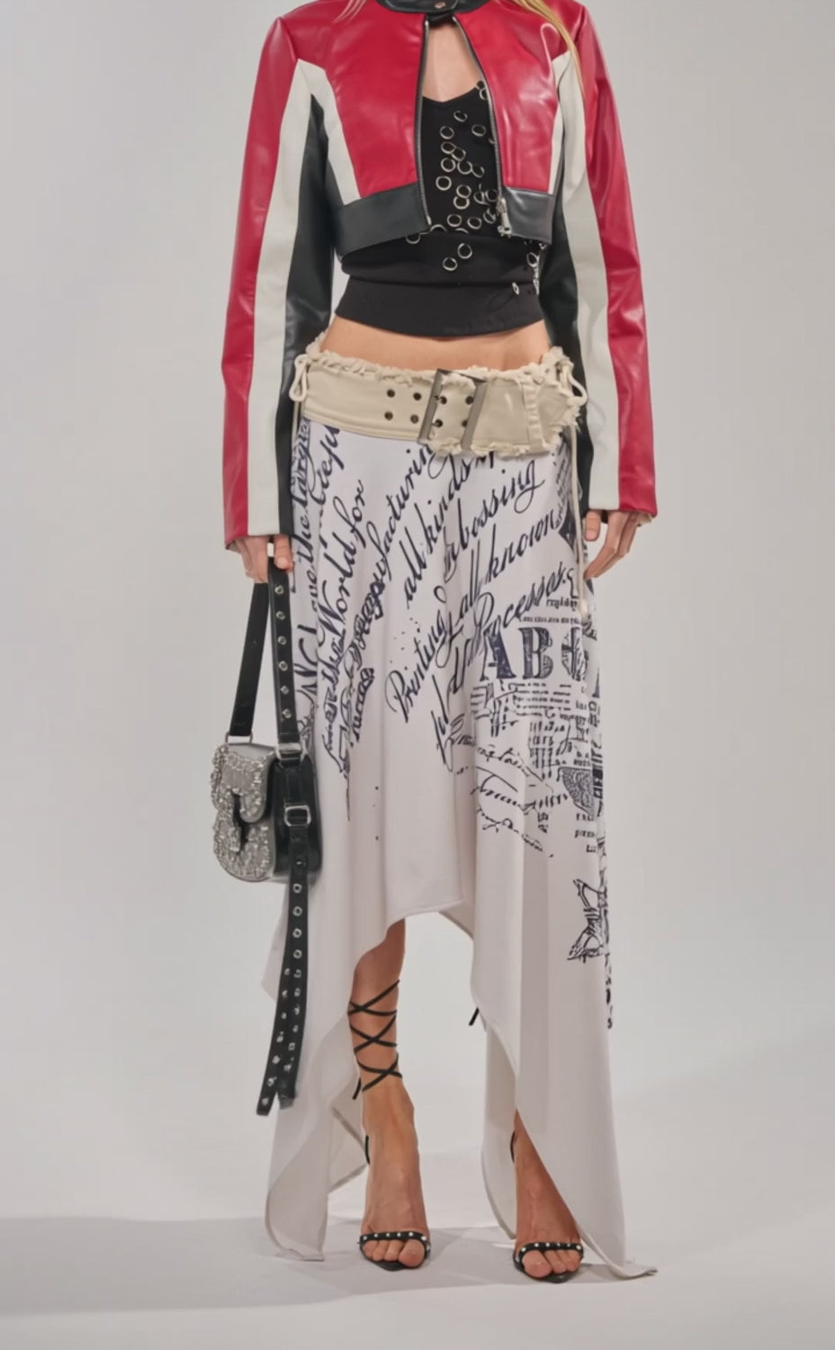 Video of woman modeling the ON YOUR MARKS CROPPED MOTO jacket, a studded black bag, black top, and a cream skirt with black cursive writing as she poses against a plain light background.