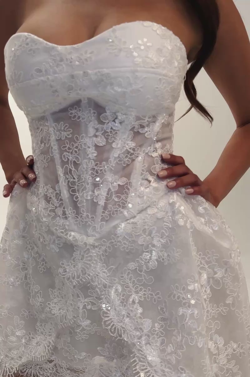 A video of a woman wearing the BACHELORETTE LACE MINI DRESS IN IVORY—a white strapless mini dress with a fitted bodice, flared skirt, and delicate lace detailing, perfect for a bridal party look. 