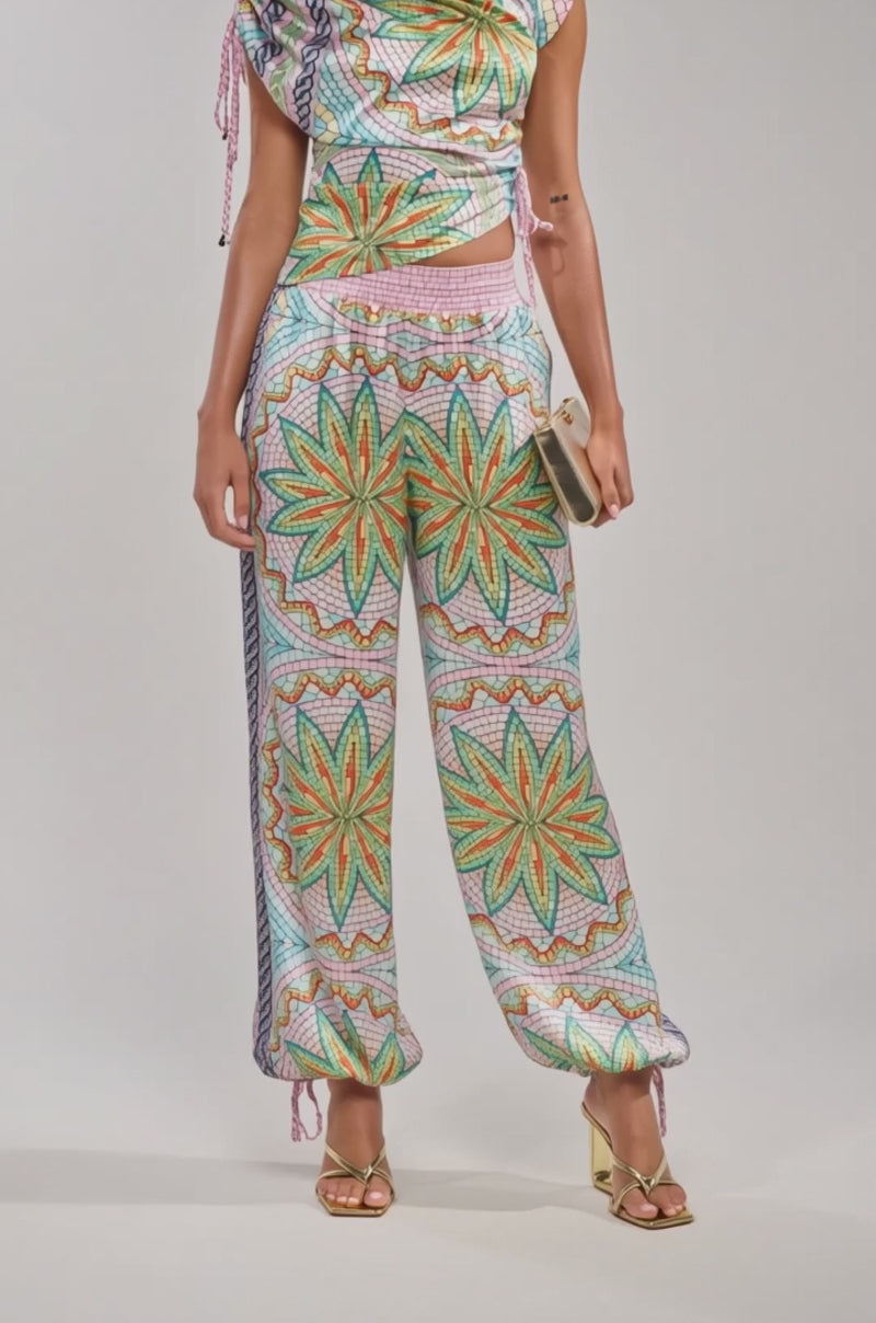 A video of a model wearing  the ALL DIFFERENT WAYS SATIN PALAZZO PANT—colorful wide-leg pants with starburst designs—paired with gold heels and a beige clutch.