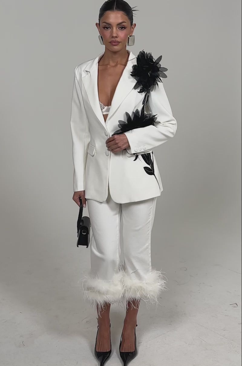 Video of woman modeling the TRIPPY LIKE ME DENIM PANT WITH FEATHER, a white jacket, black pointed-toe heels, and holding a small black handbag standing against a plain white background.