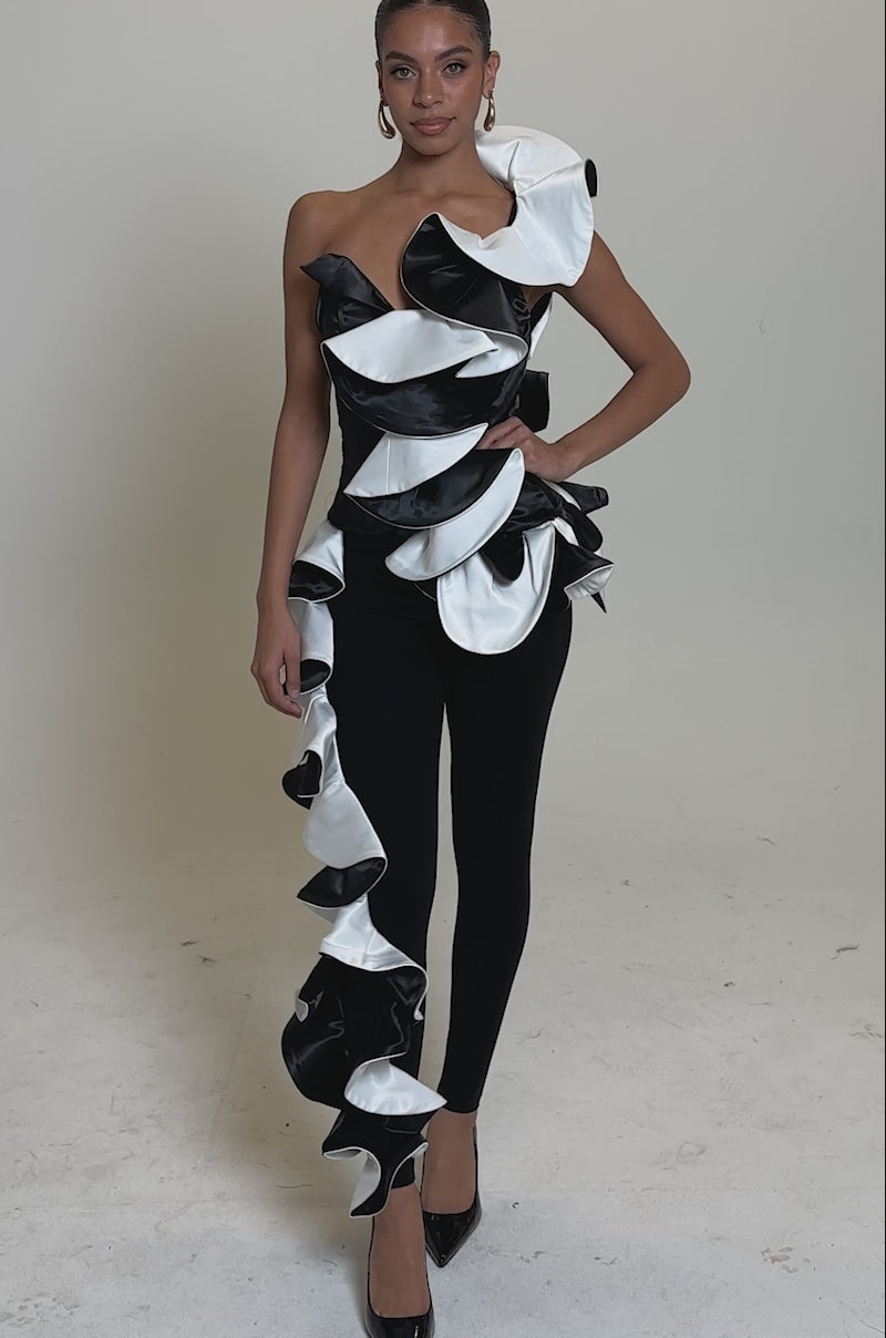 A video of a woman wearing the SNOB IS A HABIT RUFFLE DETAIL SATIN TOP, featuring dramatic black and white satin ruffles, paired with black high heels against a plain light background.