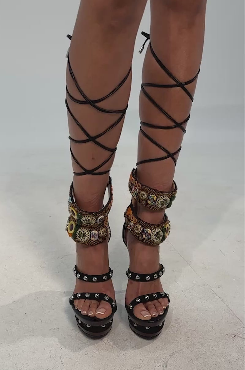 Video of woman modeling the AZALEA WANG KISMETRA BLACK BEAD EMBELLISHED PLATFORM STILETTO SANDAL, featuring studded platforms, colorful beaded straps, and black lace-up ties wrapping around the calves against a plain light gray background.