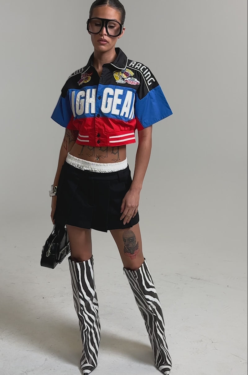Video of woman modeling the HIGH GEAR CROPPED BUTTON DOWN motorsport-inspired shirt, a pleated black mini skirt with a white waistband, and bangles, highlighting her streetwear style.