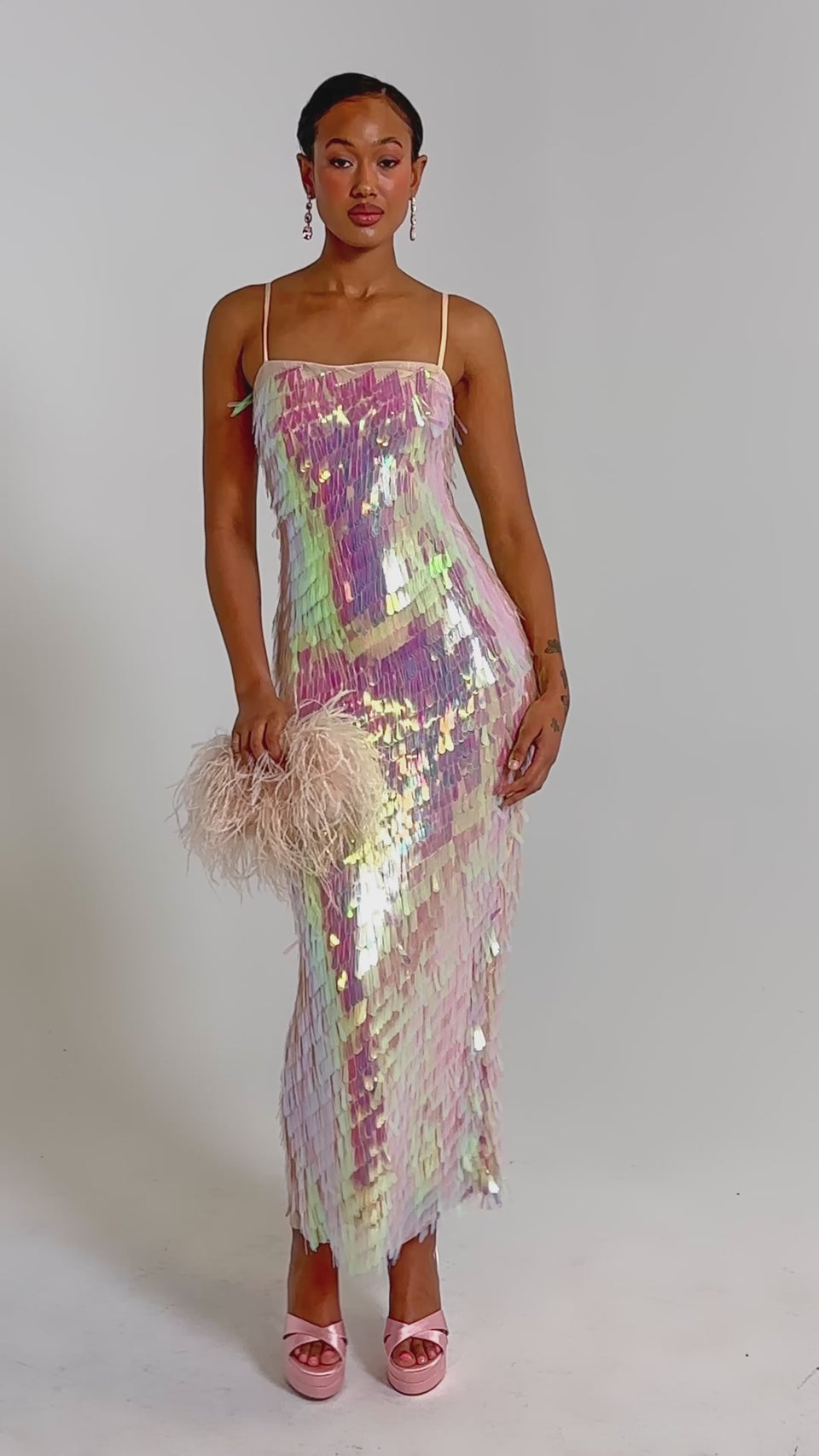 A video of a woman in the ALWAYS SHINING SEQUIN MAXI DRESS with thin straps, holding a feathered clutch. 