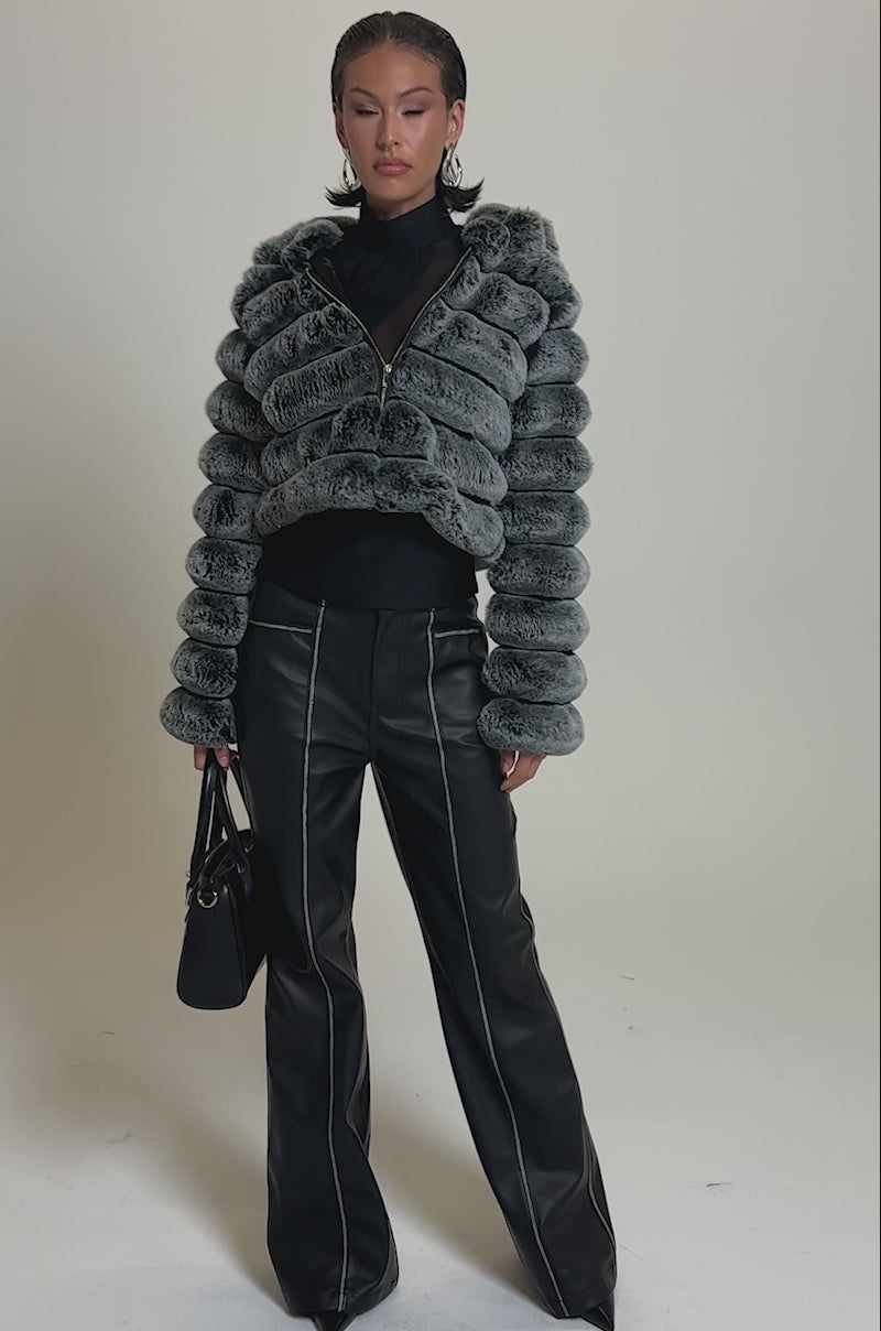 A video of a woman in the POPCORN FAUX FUR IN GREY jacket over a black turtleneck and high-waisted black leather pants with white stitching, accessorized with pointed heels and a black handbag against a plain gray background.