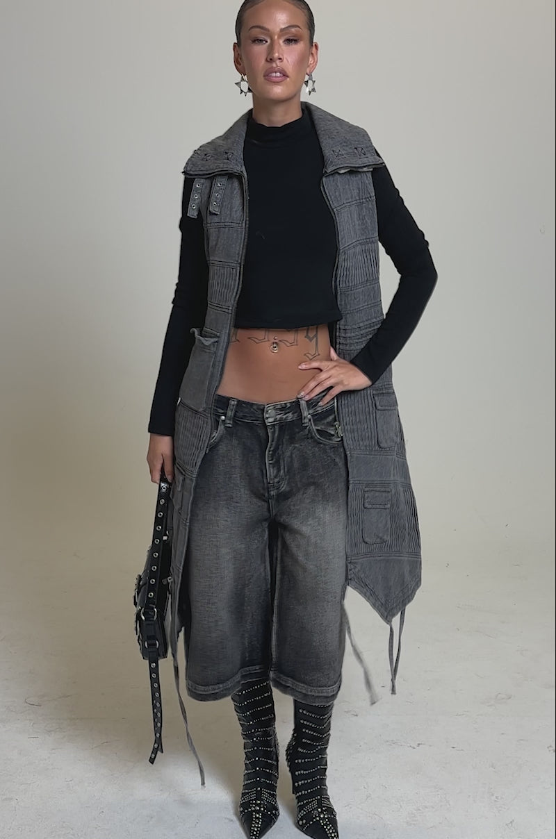 A video of a woman wearing the KEEP YOU WAITING DENIM VEST—a long, textured gray sleeveless coat with a high buckle collar—over a black top and studded boots. She holds a black handbag, her hair slicked back, against a plain light gray background.