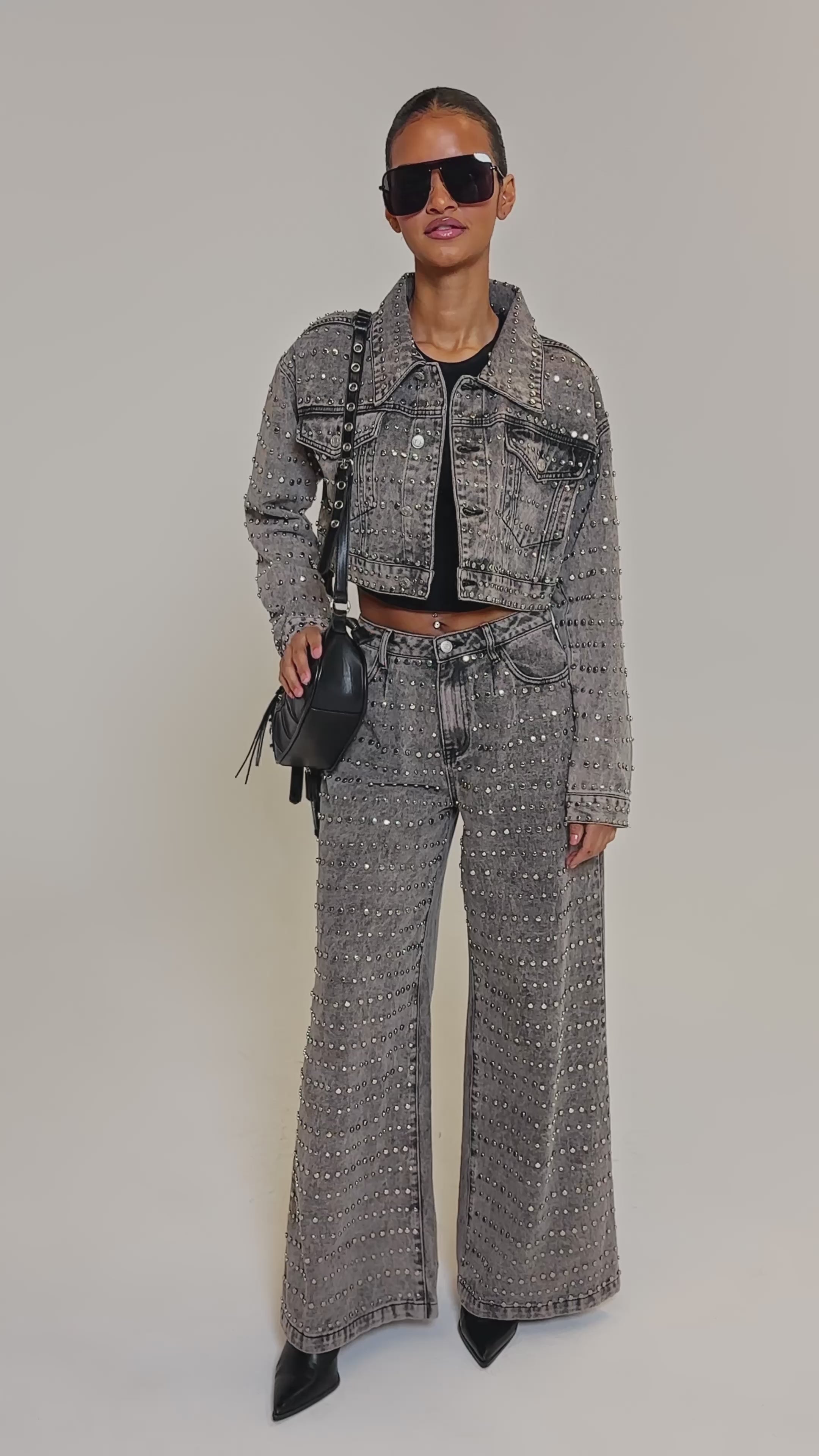 Video of woman modeling the RIZZO STUDDED DENIM JACKET with matching high-waisted pants, a black crop top, and a black shoulder bag against a plain background.