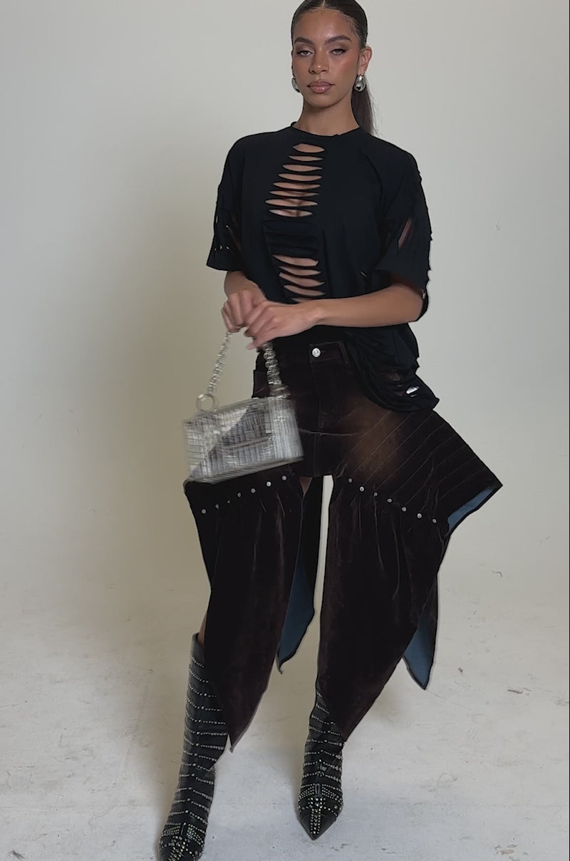 A video of a woman in the SLASHED UP OVERSIZED SHORT SLEEVE TEE IN BLACK, paired with a brown asymmetrical skirt, knee-high patterned boots, and a small geometric handbag against a plain light background.