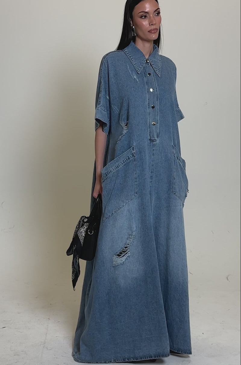 A video of a woman in THE BEST OVERSIZED DENIM JUMPSUIT—floor-length, relaxed fit, distressed denim with large front pockets and short sleeves. She holds a black handbag and wears heeled shoes.