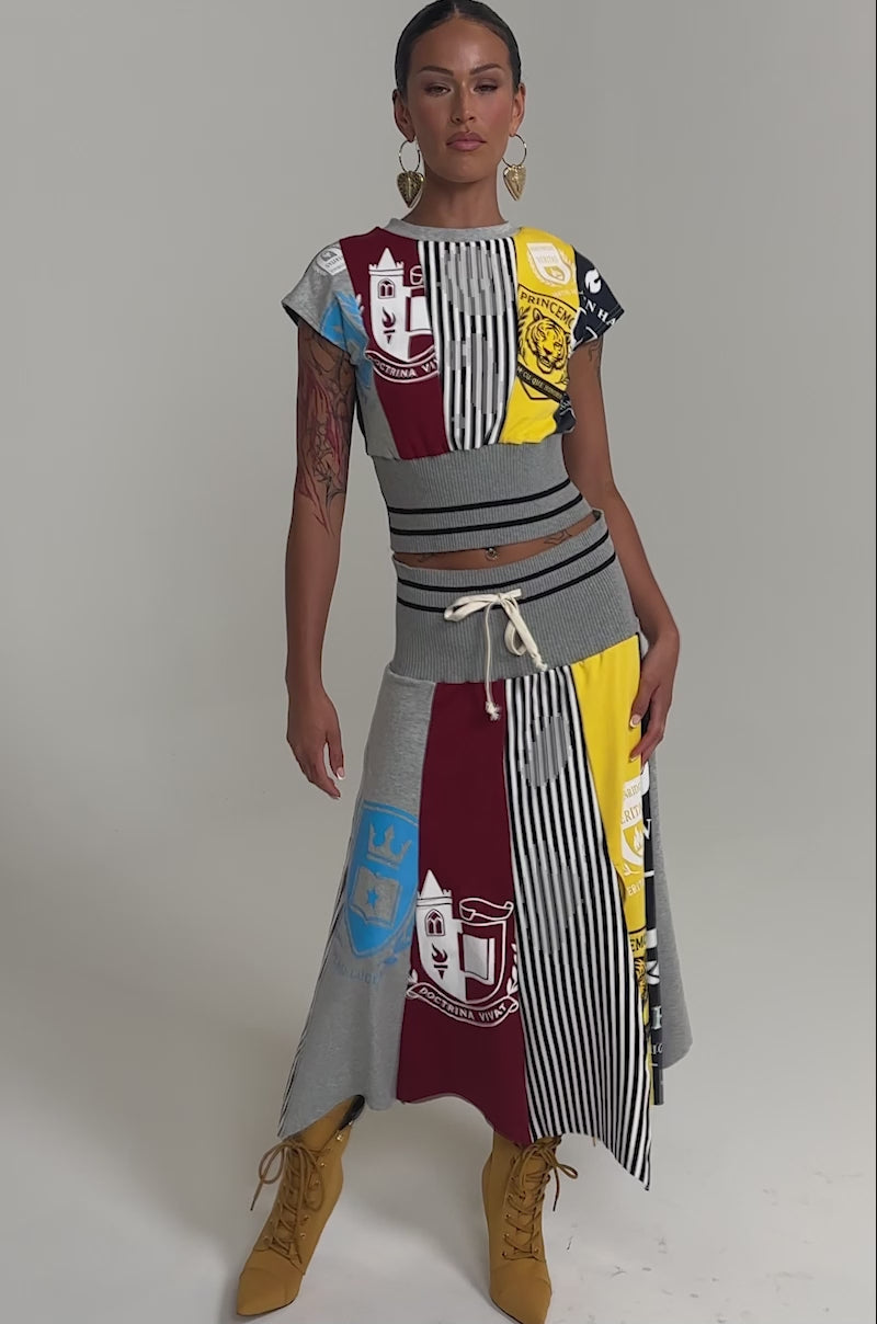 Video of woman modeling the MIXED FEELINGS MIDI SKIRT—a colorful, sporty patchwork piece with crests, stripes, and text—paired with a sleeveless top, yellow lace-up boots, and large hoop earrings against a plain background.