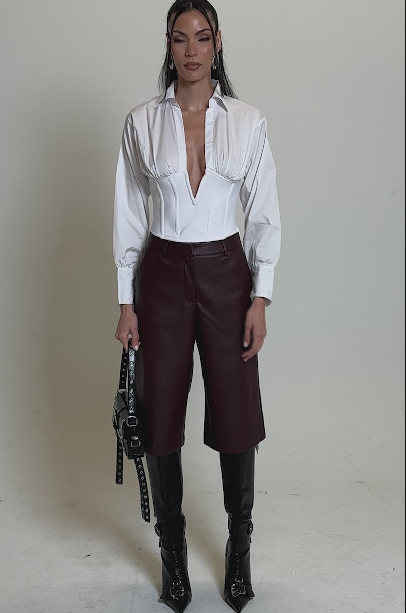 Video of woman modeling the BEST ONE YET BERMUDA SHORT in burgundy, black tall boots, and a black handbag. Her long dark hair is styled back and she wears statement earrings.