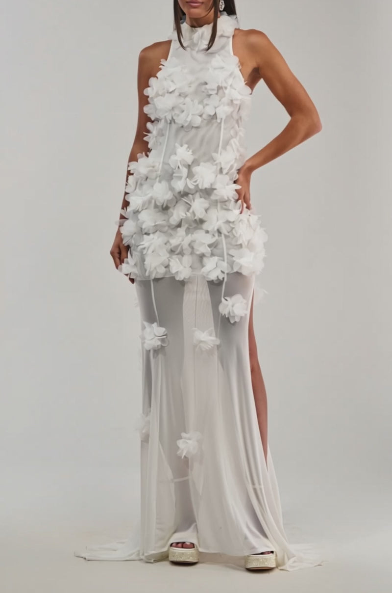 A video of a woman wearing the PARTY FAVOR RUFFLE MAXI DRESS IN WHITE, a sleeveless formal maxi dress featuring 3D floral appliqués on the upper layer and a sheer skirt. 