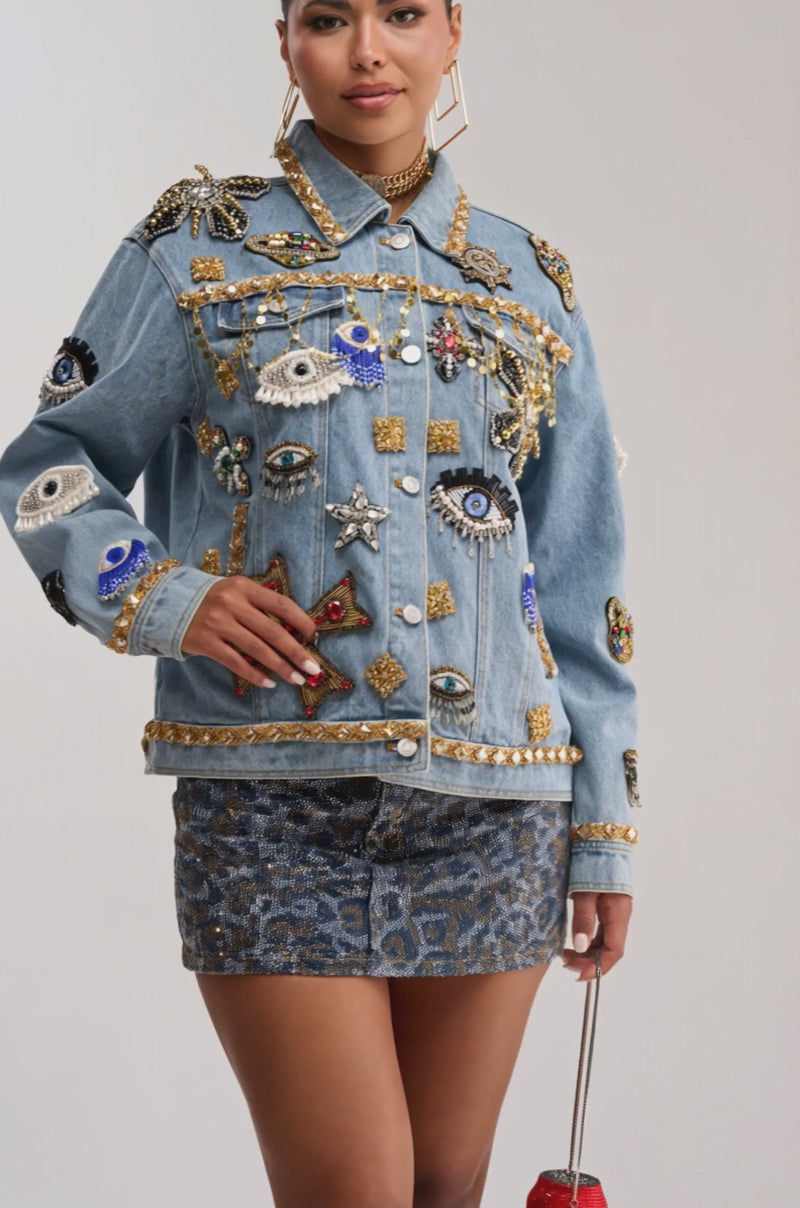 Video of woman modeling the IN PLAIN SIGHT EMBELLISHED DENIM JACKET adorned with embroidered beading, eyes, stars, and gold accents, paired with a black crop top and a patterned mini skirt.