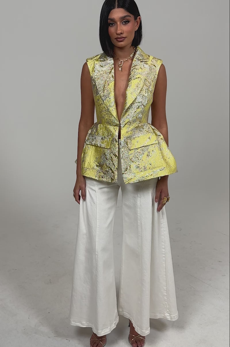 Video of woman modeling the NAOMI BLAZER VEST IN BROCADE, a yellow metallic sleeveless vest with a deep V neckline, peplum hem, and front pockets, paired with cream pants against a plain background.