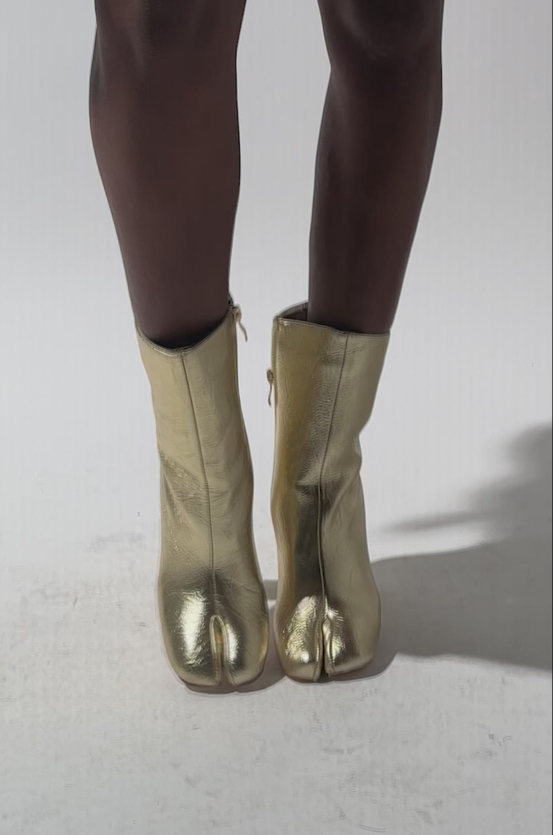A video of a person wearing a metallic, patterned skirt with AZALEA WANG BILHA GOLD SPLIT TOE CHUNKY BOOTIE, standing against a plain light background. Only the lower half of the body is shown.