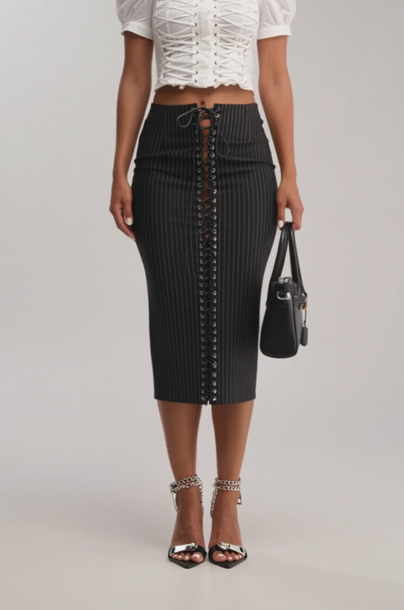 Video of woman modeling the LIVI STRETCHY PINSTRIPE MIDI SKIRT with a white cropped top, chain-strap heels, and a black handbag—creating a chic, high-waisted date night look featuring front lace-up detail.