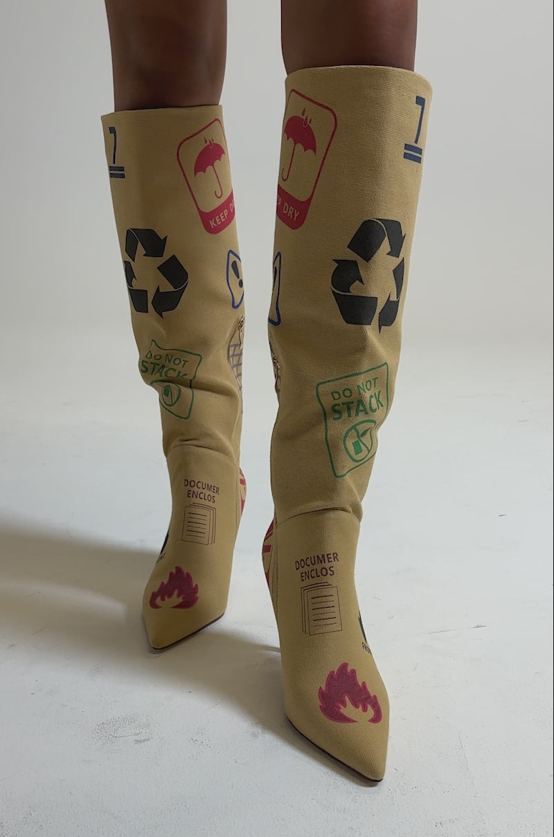 A video of a person wearing the AKIRA GOLD WHOLE PACKAGE TAN STILETTO BOOT, featuring knee-high tan color, pointed toe, high thin heel, and vibrant graphic prints like recycling and fragile icons, paired with a tan skirt.