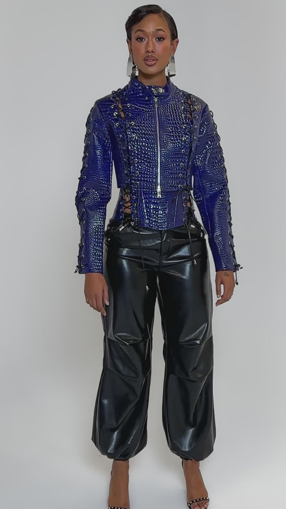 Video of woman modeling the HIT THE SWITCH EMBOSSED MOTO, a shiny blue crocodile print zip-up jacket with lace-up details, paired with black faux leather pants and bold silver earrings, set against a plain light background.
