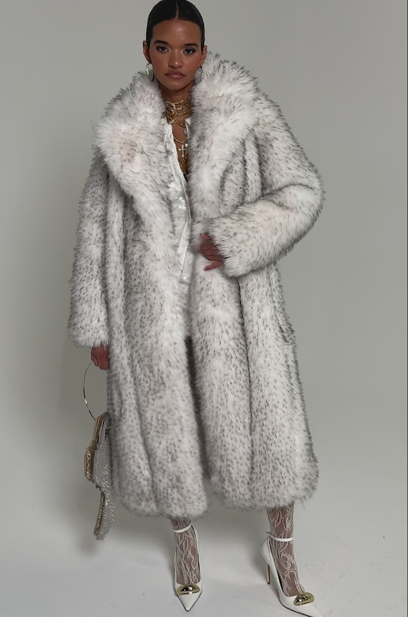 Video of woman modeling the HEAVY ON THE APRES FAUX FUR COAT over a white outfit. They hold a rhinestone key-shaped handbag and wear gold heels with white patterned tights.