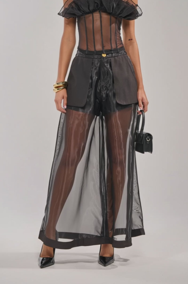 Video of woman modeling the FAN OUT SHEER CORSET, an off-the-shoulder sheer black top with ruffles, paired with matching high-waisted pants and gold accessories—a chic look for date night.