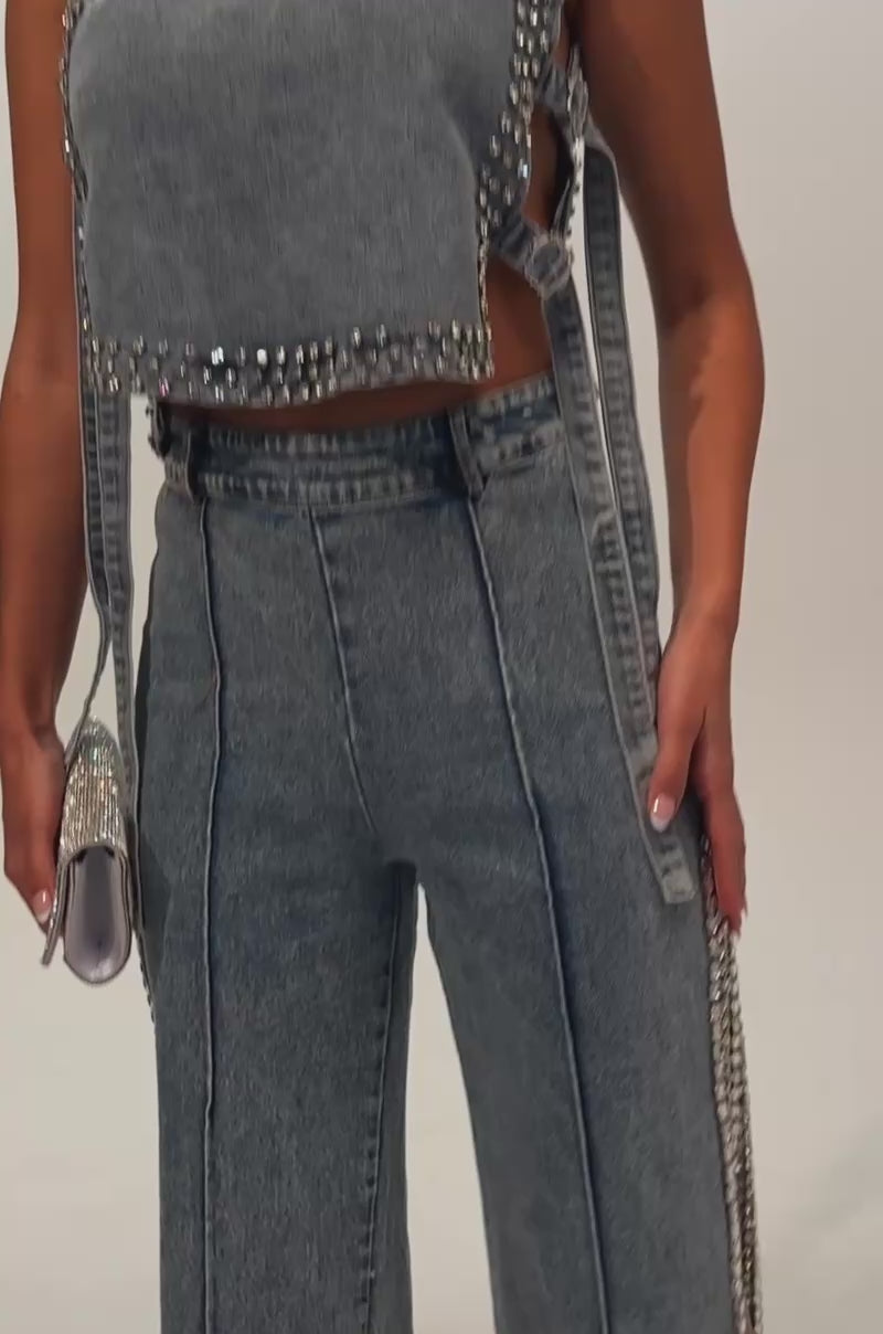 A video of a model wearing the POPPING OUT RHINESTONE DENIM PANT—high-waisted, straight-leg jeans in a blue wash featuring front seams and bold high side slits embellished with rhinestones.