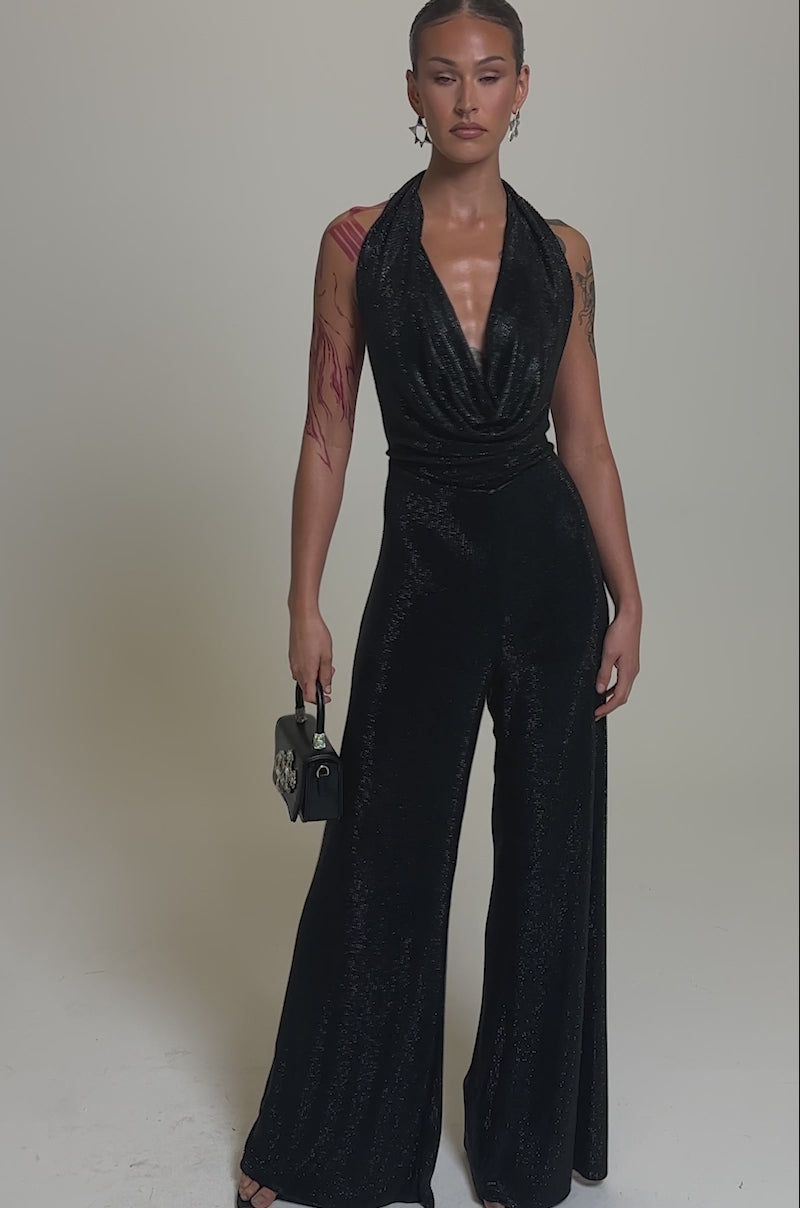 Video of woman modeling the MAKE YOU DO A DOUBLE TAKE RHINESTONE JUMPSUIT IN BLACK with a draped neckline, holding a black handbag. She has slicked-back hair, bold earrings, and a red tattoo sleeve on her right arm.
