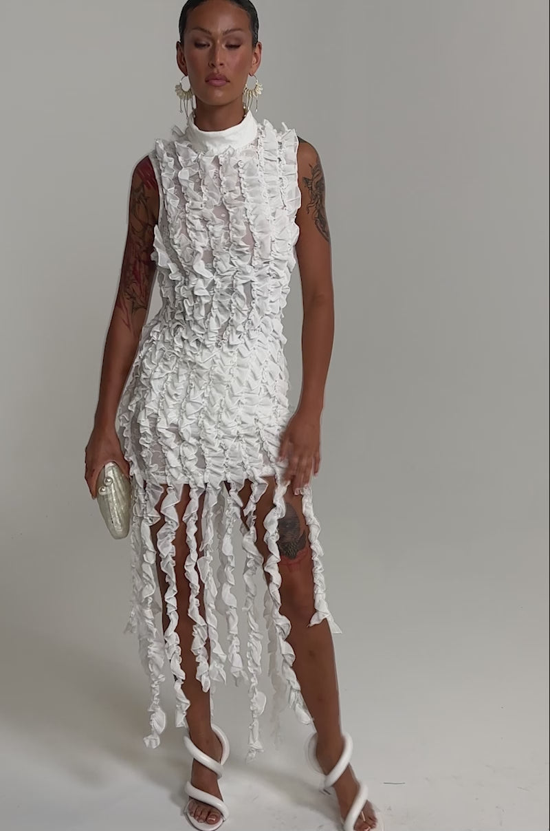 A video of a woman wearing the FOR THE FRILL OF IT MINI DRESS IN WHITE, paired with white heeled sandals, large earrings, and holding a small oval clutch.