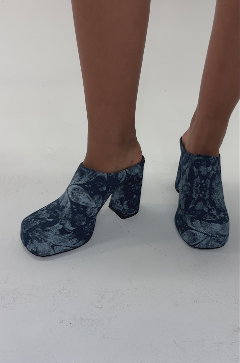 Video of woman modeling the AZALEA WANG FLOREA DENIM PRINT MULE—a high-heeled, blue patterned mule with a block heel and closed toe—set against a plain, light background.
