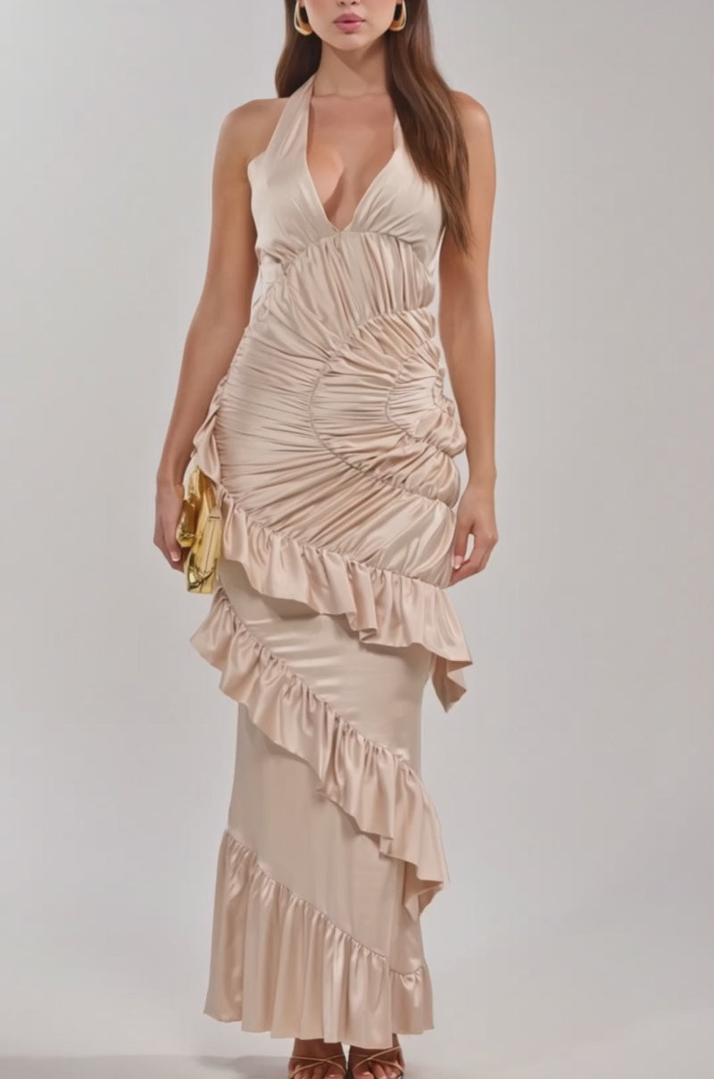 A video of woman in the MAKE YOU MINE RUFFLE SATIN MAXI DRESS, featuring cream satin with ruffles, ruching, and a halter neck, stands against a plain background holding a small gold clutch and wearing high-heeled sandals.