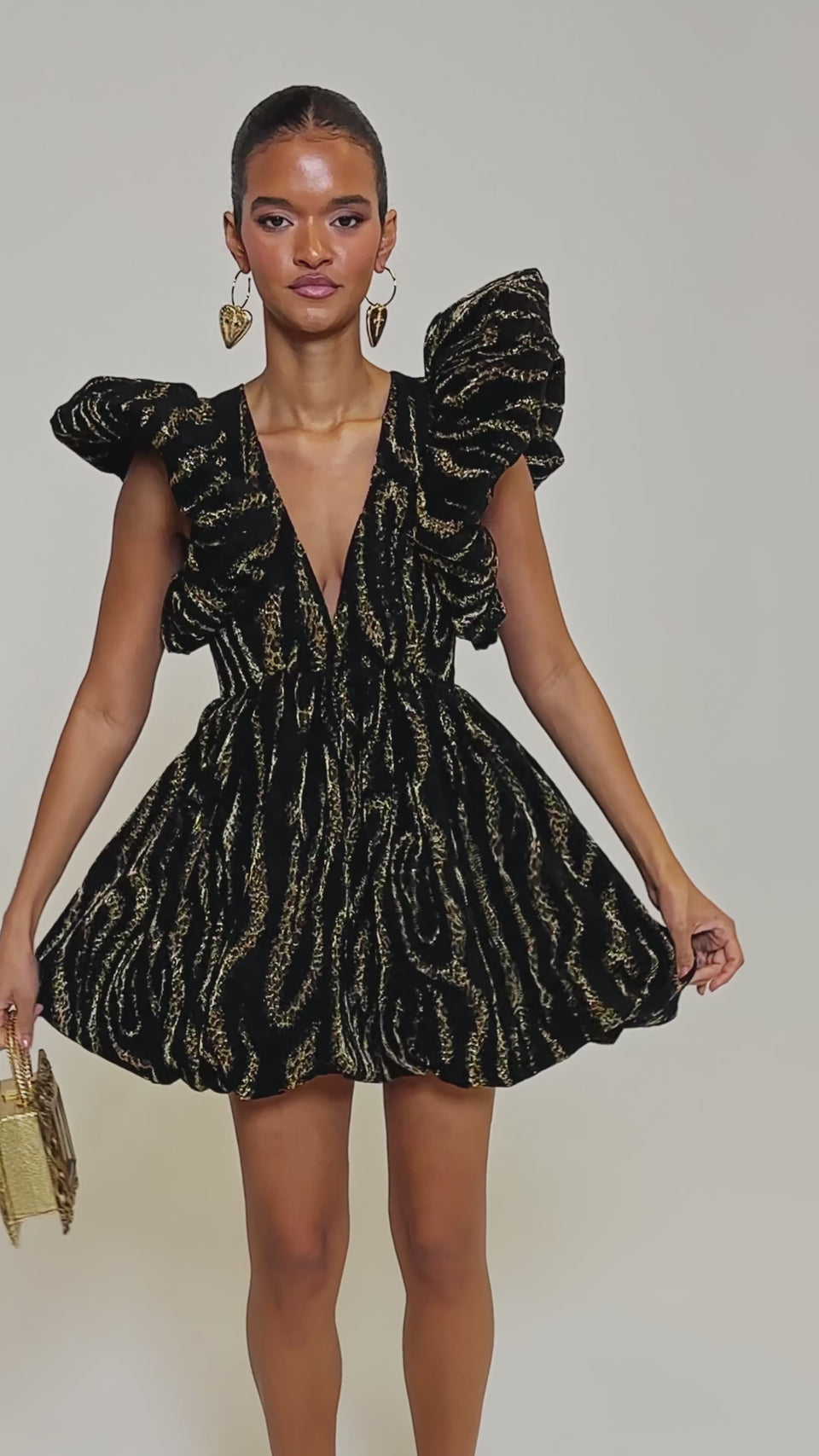 Video of woman modeling the WILD HEART BROCADE BUBBLE MINI DRESS, featuring black and gold brocade, puffed sleeves, a deep V-neckline, and a flared skirt—ideal for holiday parties. She pairs it with large heart-shaped earrings and a small bag.