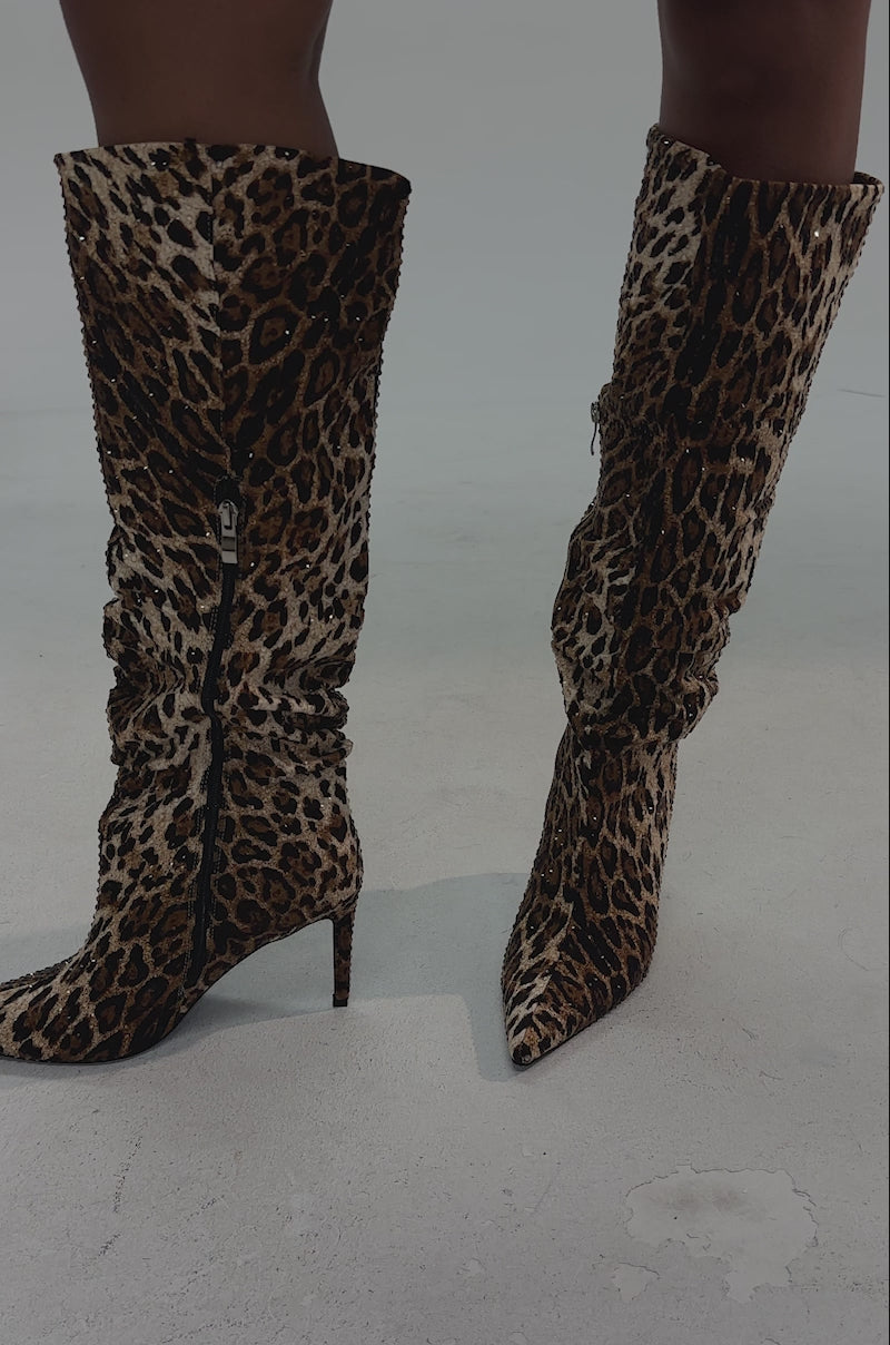 A video of a person wearing a short, fitted leopard-print skirt and the AZALEA WANG GRANT RHINESTONE LEOPARD SLOUCH BOOT with stiletto heels, both in a bold animal pattern, set against a dark background.