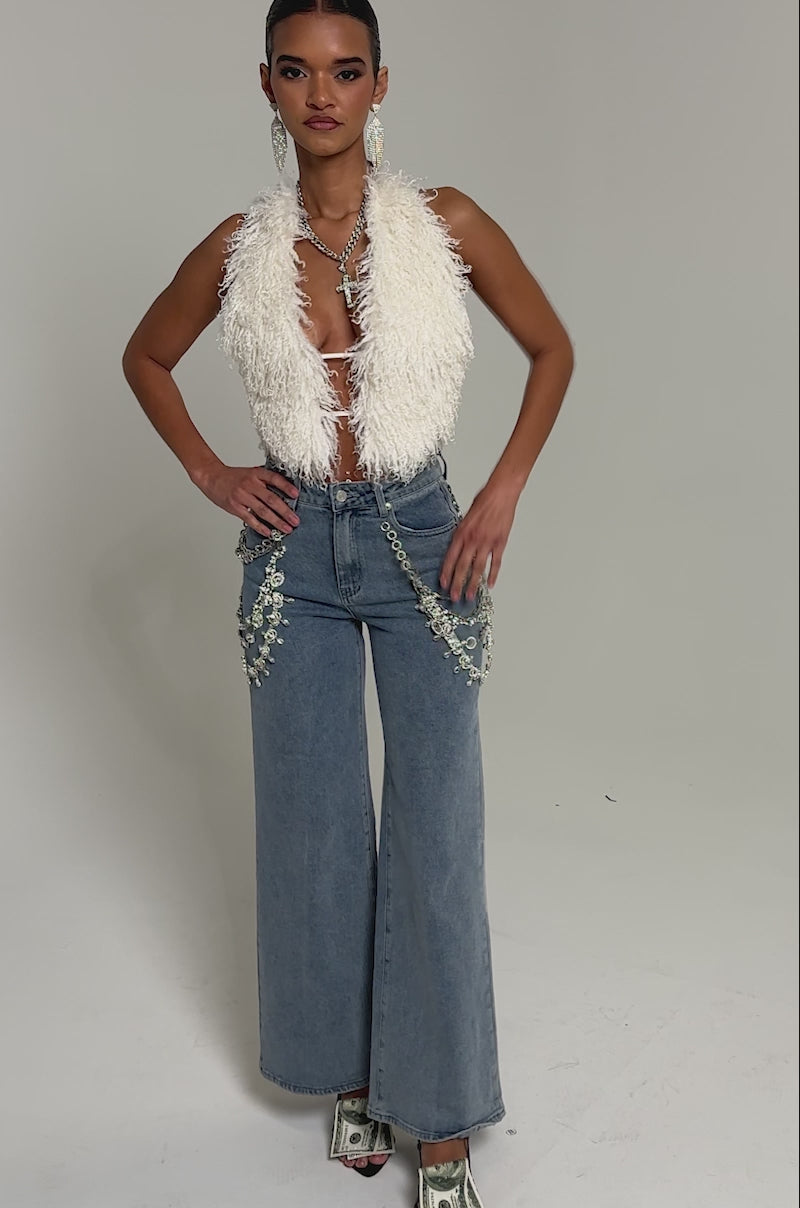 Video of woman modeling the ACT UP FUR BLOUSE IN WHITE, statement earrings, layered silver necklaces, and blue jeans with silver chain details. With slicked-back hair, she stands against a plain background.