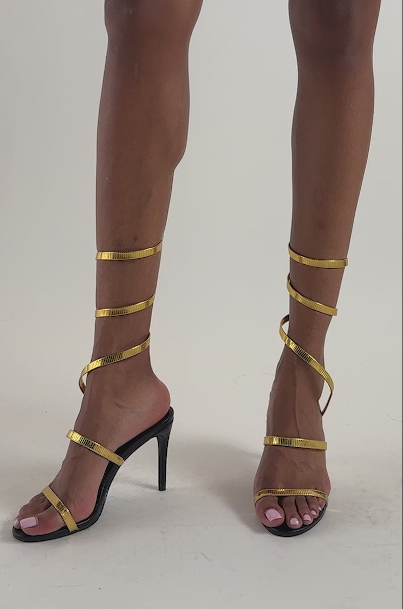 Video of a woman modeling the AKIRA GOLD STHENO GOLD METAL COIL SANDAL—elegant gold stiletto sandals with coiled straps that wrap around their legs.