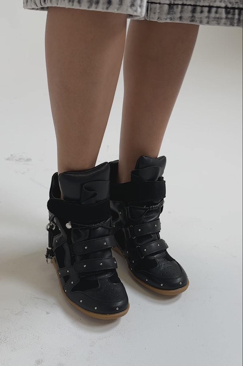 A video of a person wearing AKIRA GOLD RANGELEY black wedge sneakers with silver hardware, buckles, straps, studs, and a gum sole. 