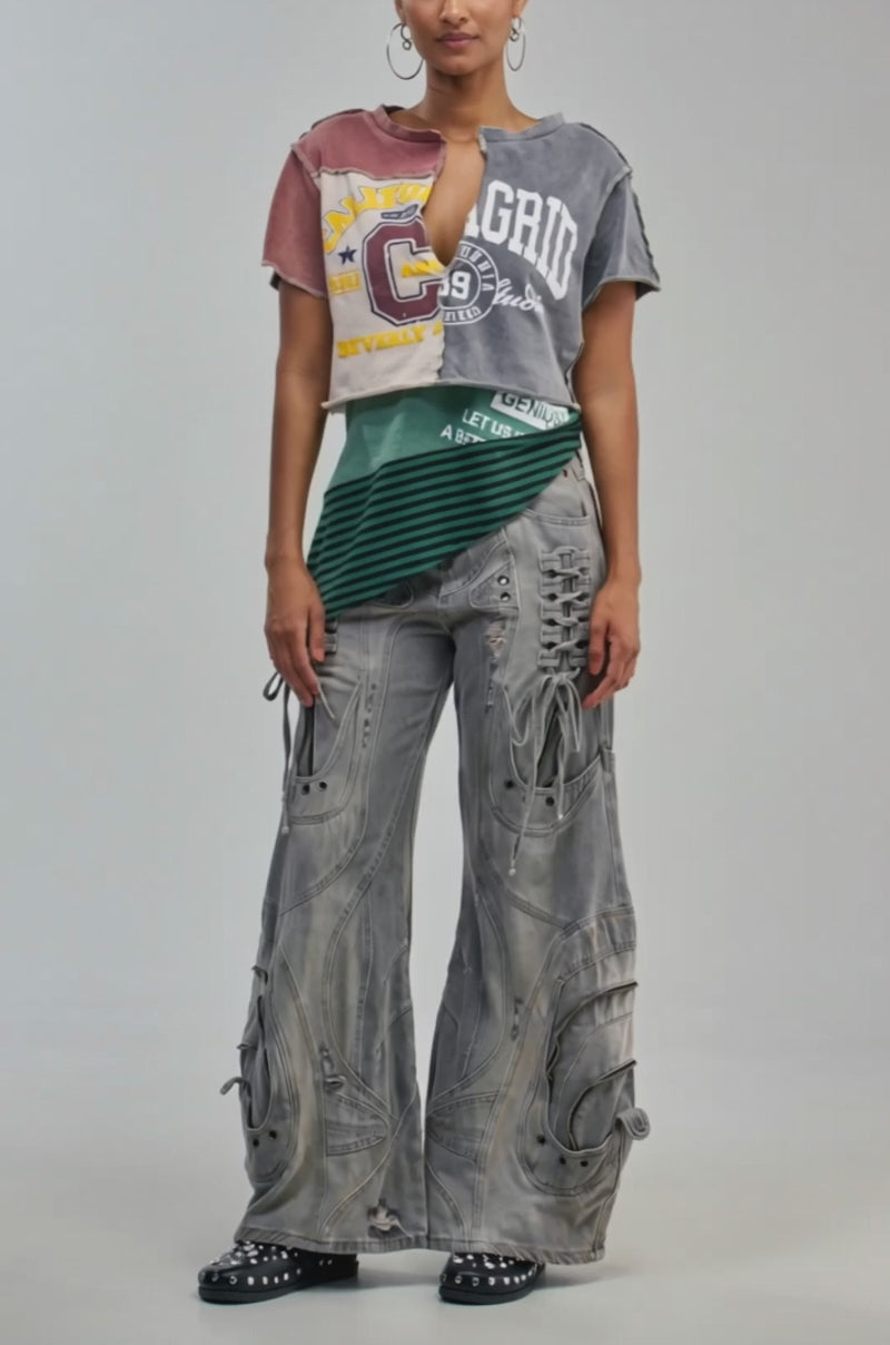 Video of a woman modeling the FIRST CLASS PATCHWORK TEE, a woman stands against a plain background. The tee features an asymmetrical patchwork design with ripped details and striped patterns. 