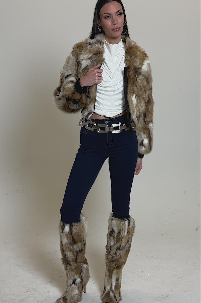 A video of a woman in the FENNEC FAUX FUR BOMBER with a cropped boxy fit, matching faux fur leg warmers, a white draped top, blue skinny jeans, hoop earrings, and a statement belt.