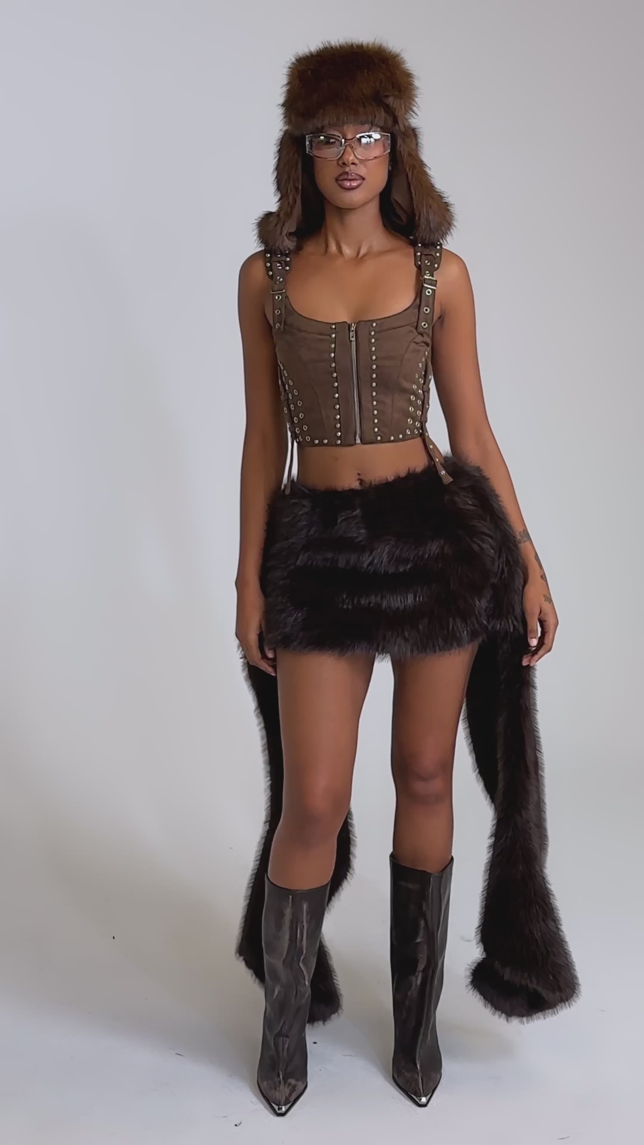 A video of a model wearing the TAKE ME TO THE RODEO HOODED FUR TOP, a person stands against a plain light background, styled with a brown fur hat, faux fur skirt with long panels, and tall black boots.
