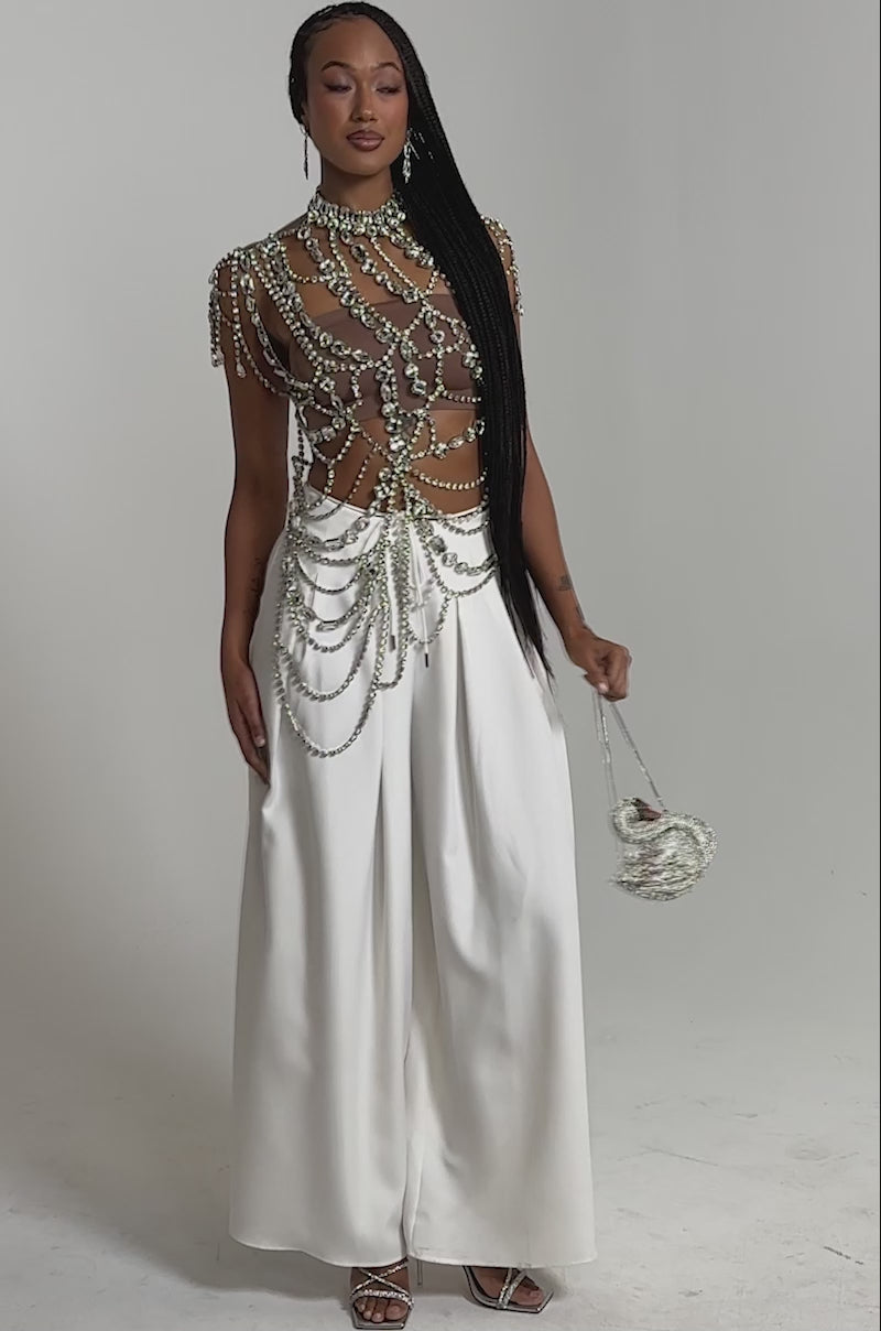 A video of a woman wearing the REGAL RHINESTONE CHAIN TOP with pearl chains, wide-legged white pants, and heeled sandals. 
